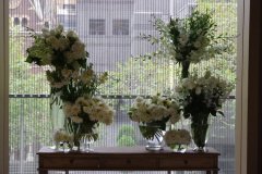 Elegant white floral arrangements displayed on a wooden table, featuring various vases and greenery, set against a backdrop of city views, ideal for wedding inspiration by Antaeus Flowers.