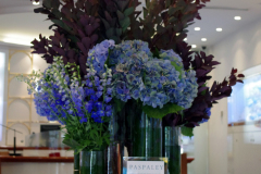 Floral arrangement featuring blue hydrangeas, purple foliage, and green accents in glass vases, showcasing corporate floral design by Antaeus Flowers.